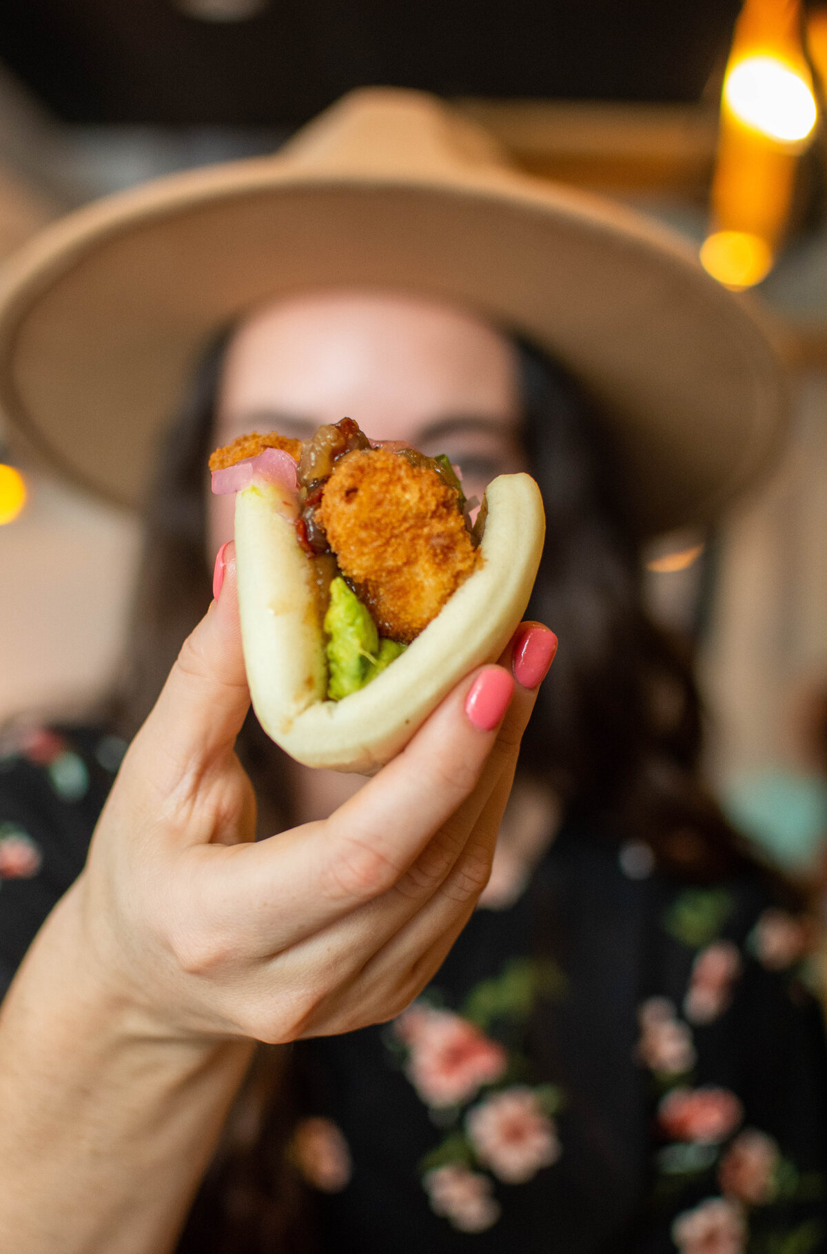 Woman with dark hair wearing a hat holding a bao in front of her face