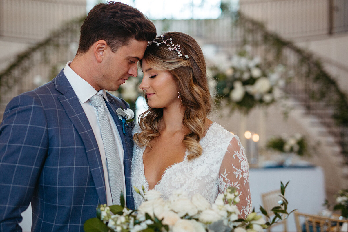 A bride and groom in a blue suit and white wedding gown pose in a banquet room with white floral arrangements and candles.