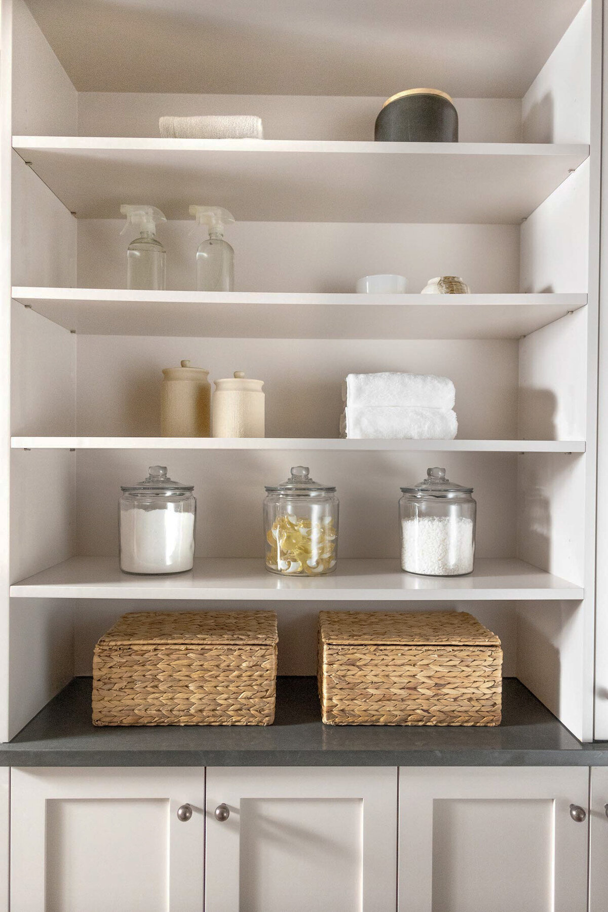 Beige cabinet with doors with antique bronze knobs. Grey quartz countertops above with open shelving painted beige. Shelves adorned with glass jars of laundry detergent and pods, cleaning spray bottles, towels, jars, and wicker baskets.