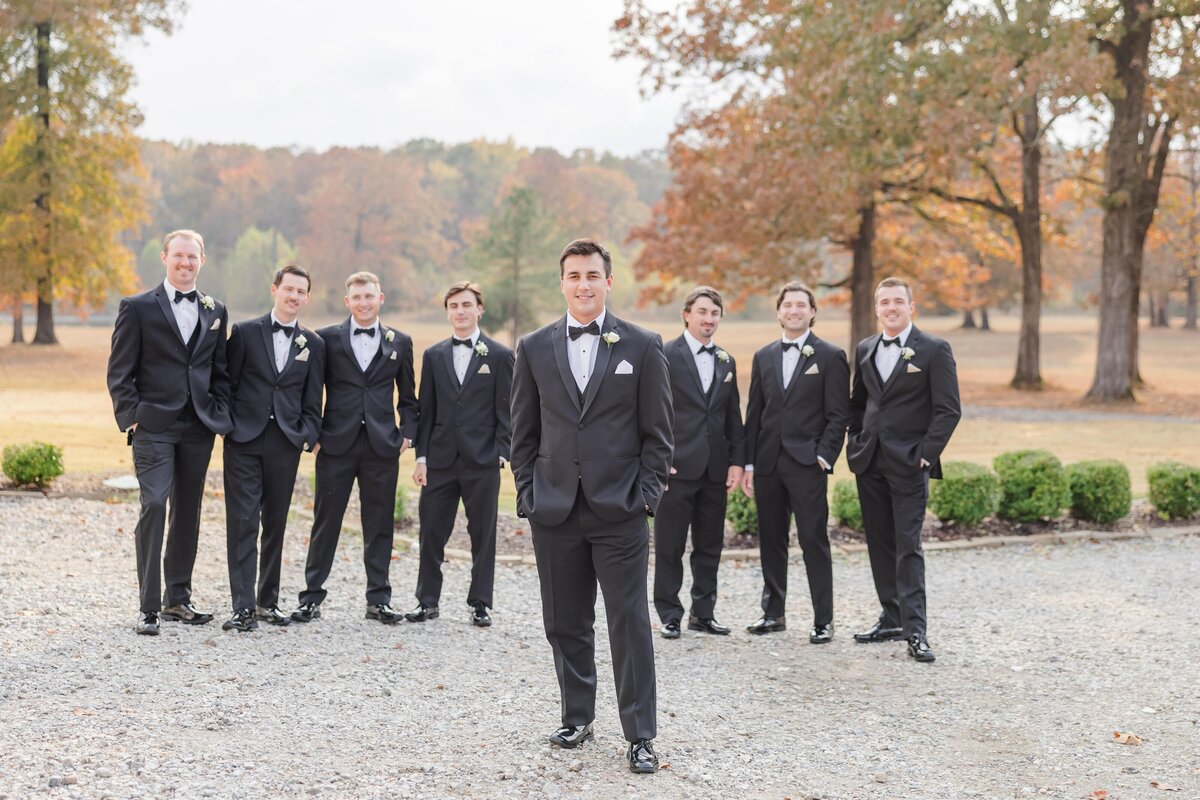 A groom stands in front of his groomsmen all in black suits with hands in pockets in gravel