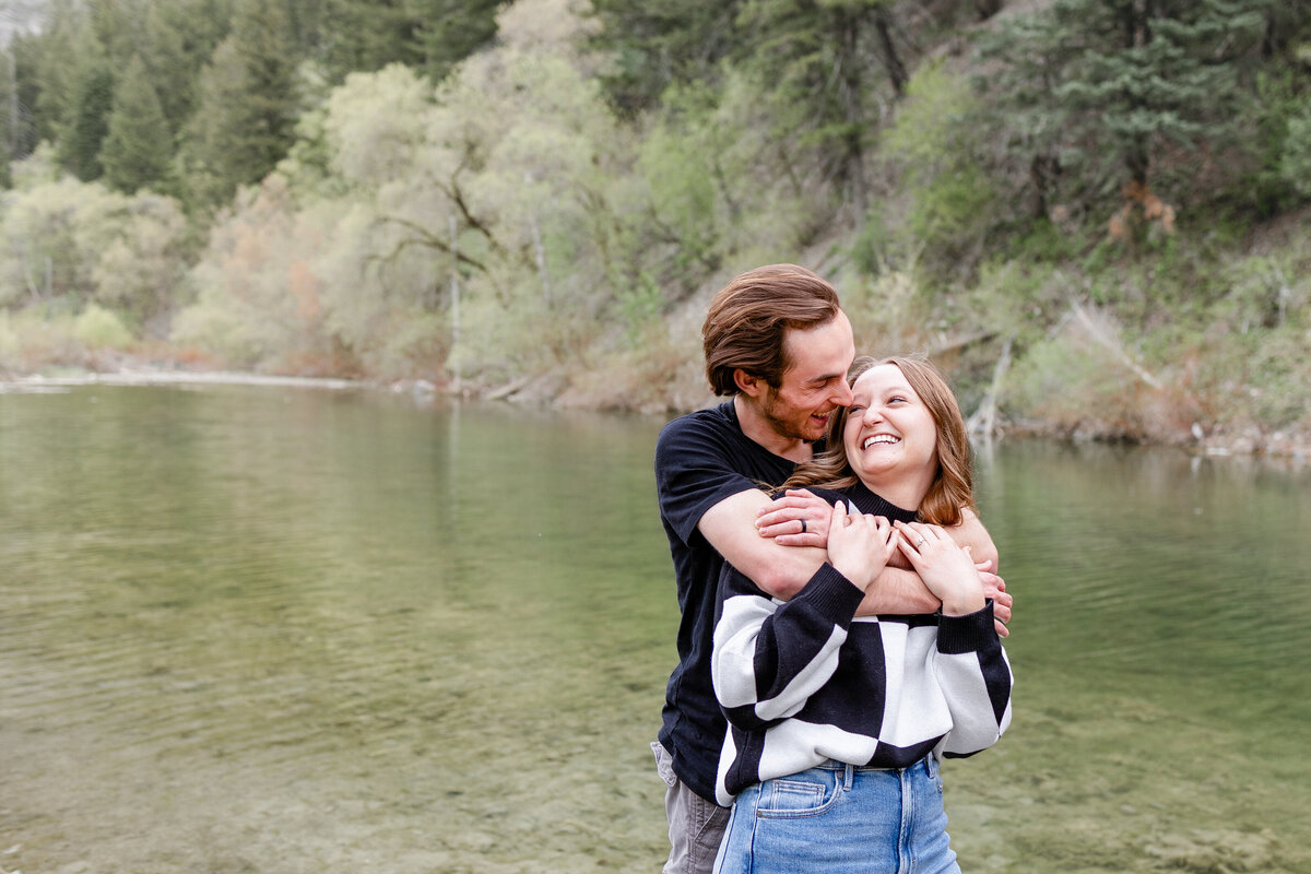 A smiling couple standing close together near the river, with the fiancé wrapping his arms around his future bride as they pose for their engagement photos.
