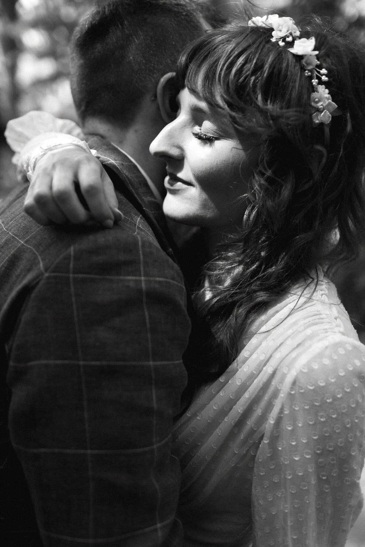 In a black and white photo, a woman in a white dress embraces a man in a suit, her eyes closed and a floral headband in her hair. Captured at Queen City Yacht Club by Toronto wedding photographer, Jennifer van Son.