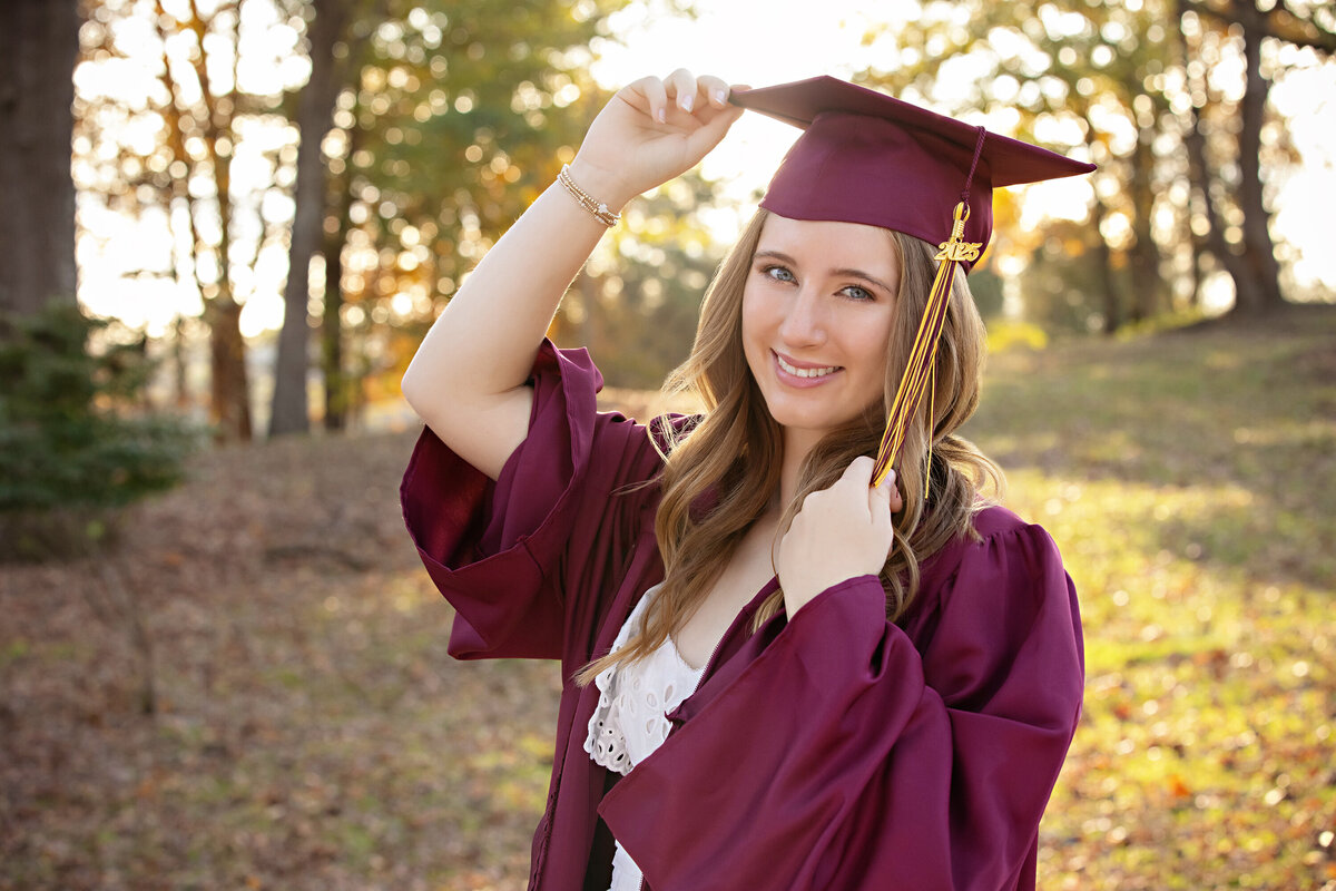 High school senior in cap and gown smiling near gravel road and autumn trees