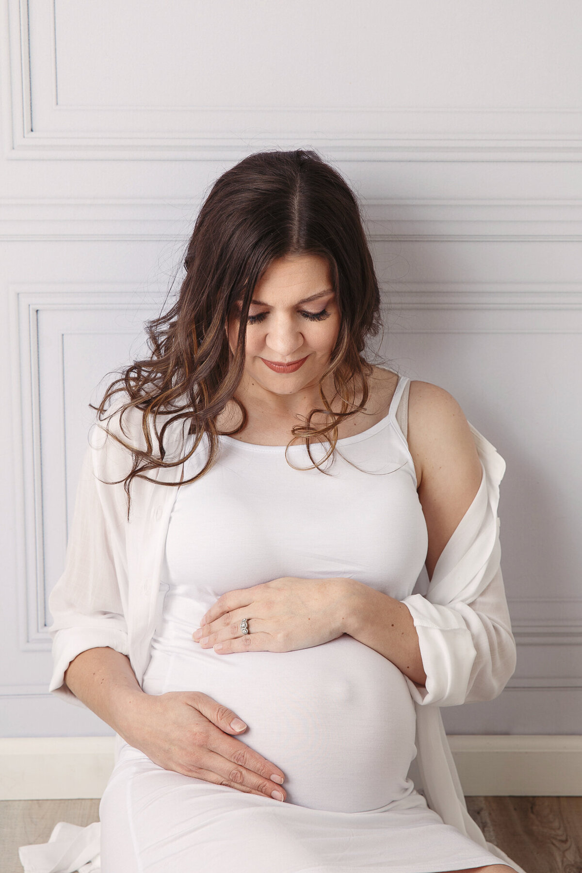 Motherhood portrait of a young pregnant woman sitting against a wall, cradling her belly and looking down at her baby bump