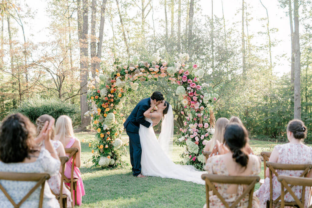 Bride and groom kiss under a colorful floral arch during romantic outdoor wedding ceremony.