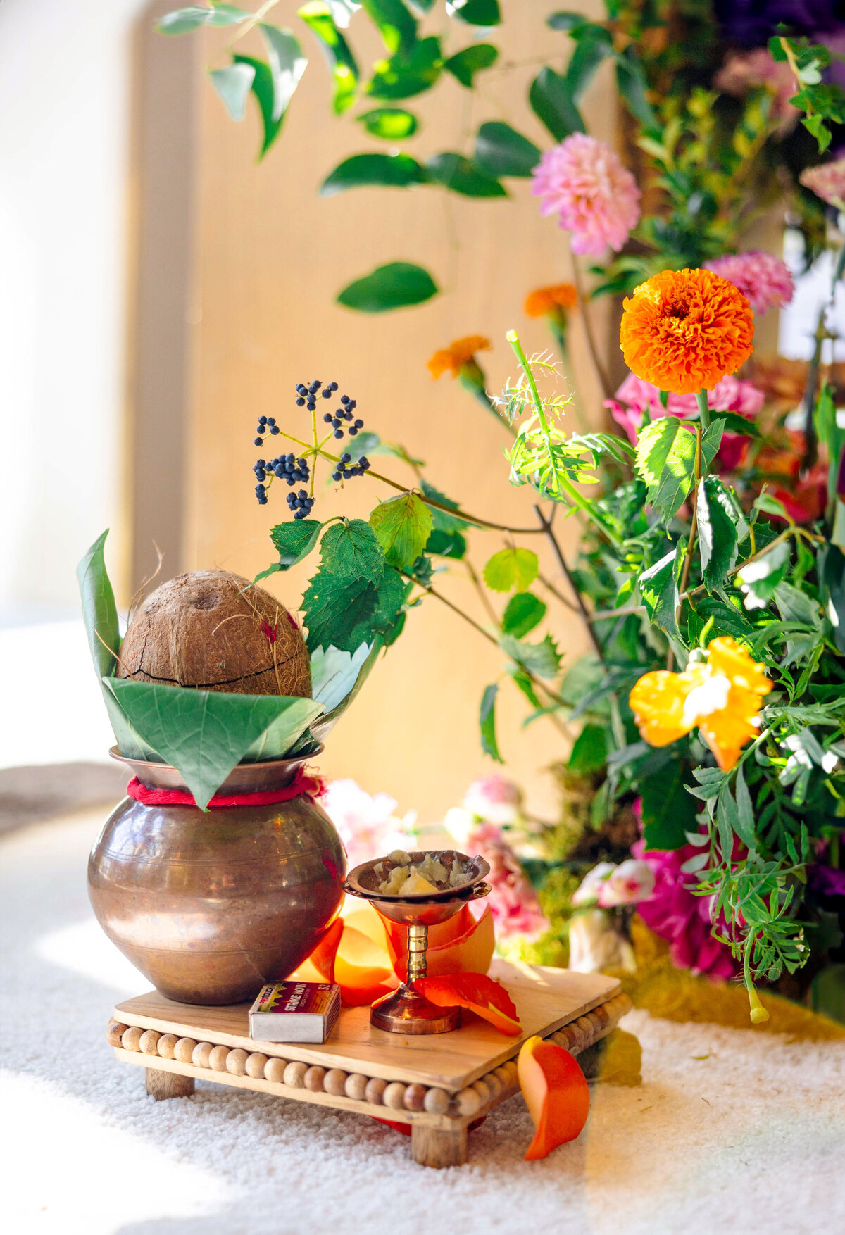 A decorated brass pot with a coconut and leaves sits on a wooden platform. Beside it is a small brass cup with flowers and a ribbon. Colorful flowers and greenery surround the setup, indicating a festive or traditional ceremony.