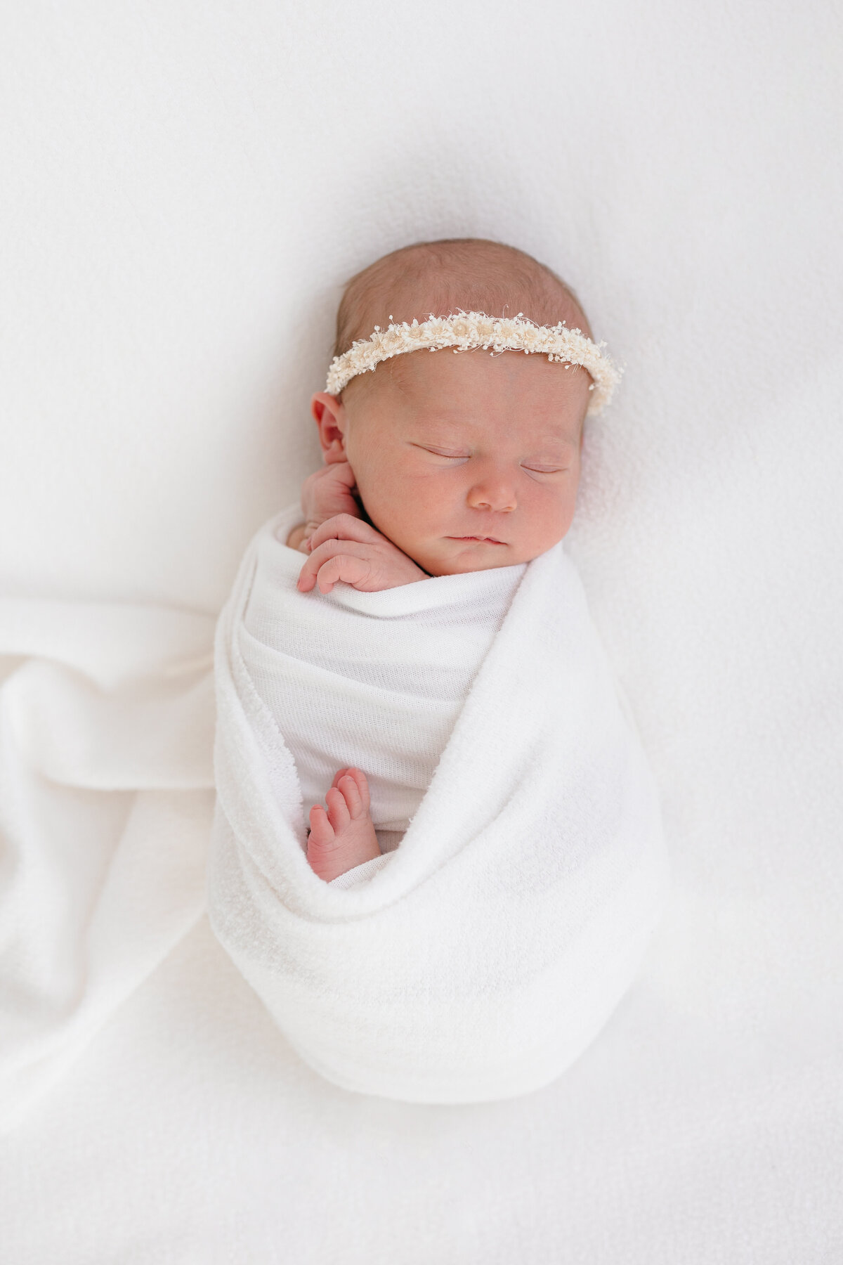 Newborn baby girl wrapped in a white swaddle with her hands gently touching her cheek