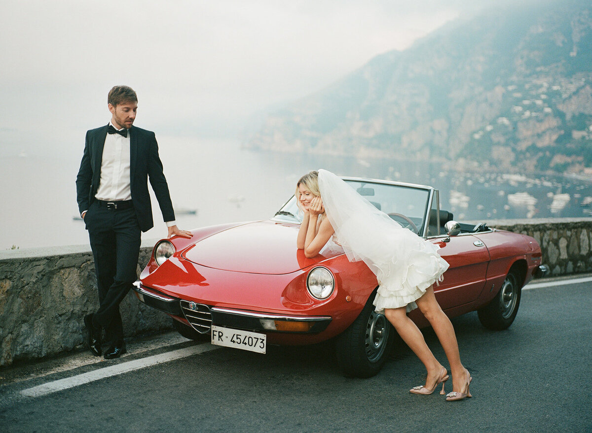 couple on the amalfi coast with a vintage car . Bride is leaning over the cars hood.