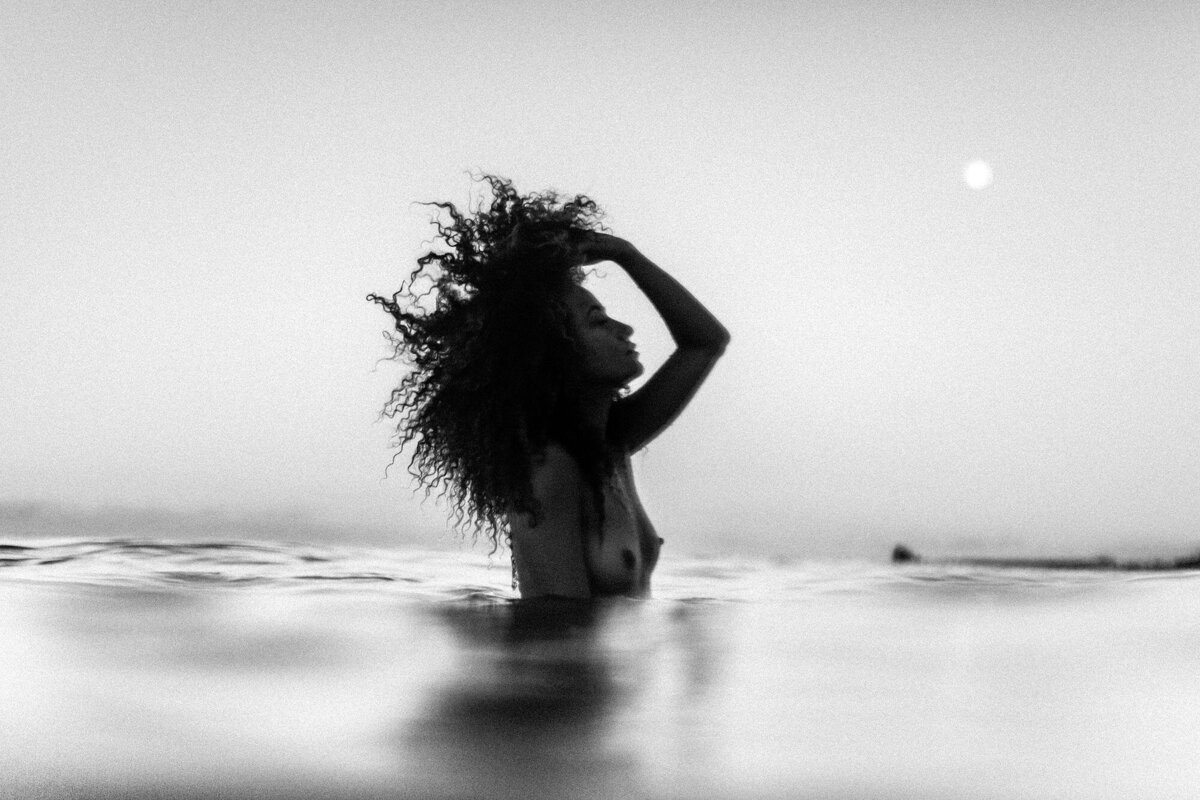 black and white photo of a woman throwing back her hair while standing in the ocean under moonlight