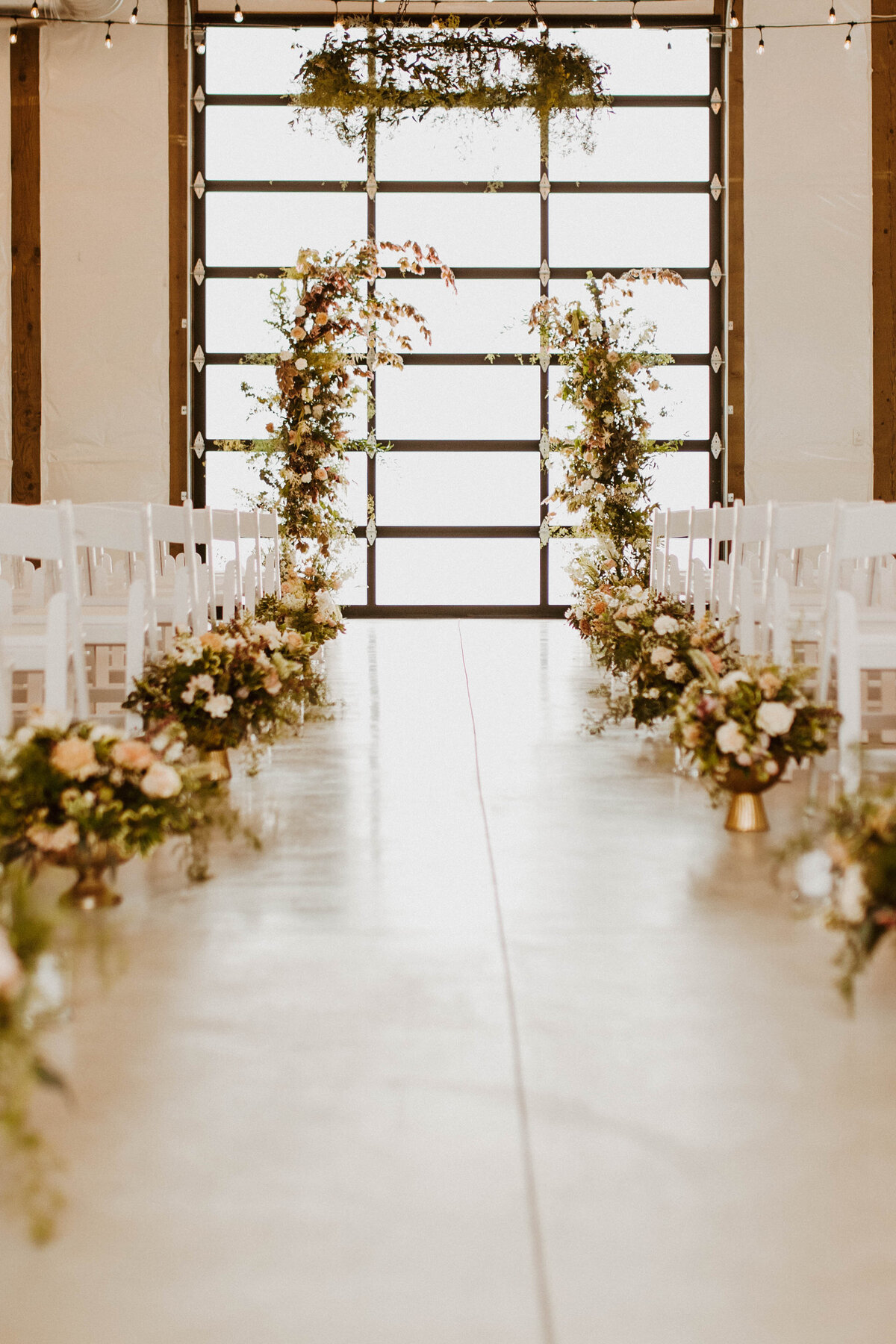 Shot of interior White Barn with rows of white chairs, flower placements at each row and a shot of the altar,  the big glass door framed with greenery and flowers.