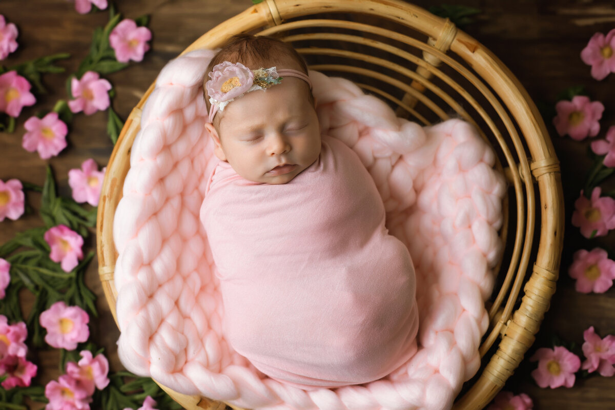 Newborn photographer, a baby girl sleeps swaddled in pink and surrounded by flowers.