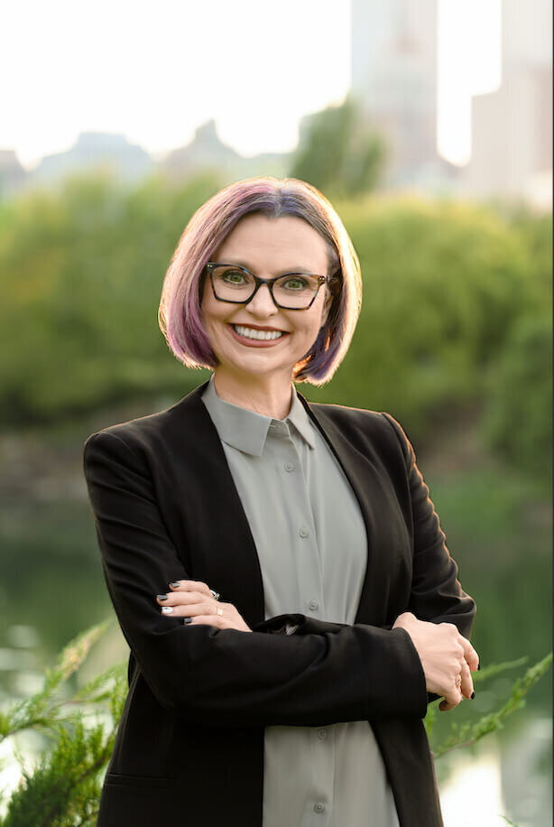 woman smiling at the camera wearing glasses in a suit with the downtown tulsa skyline in the background