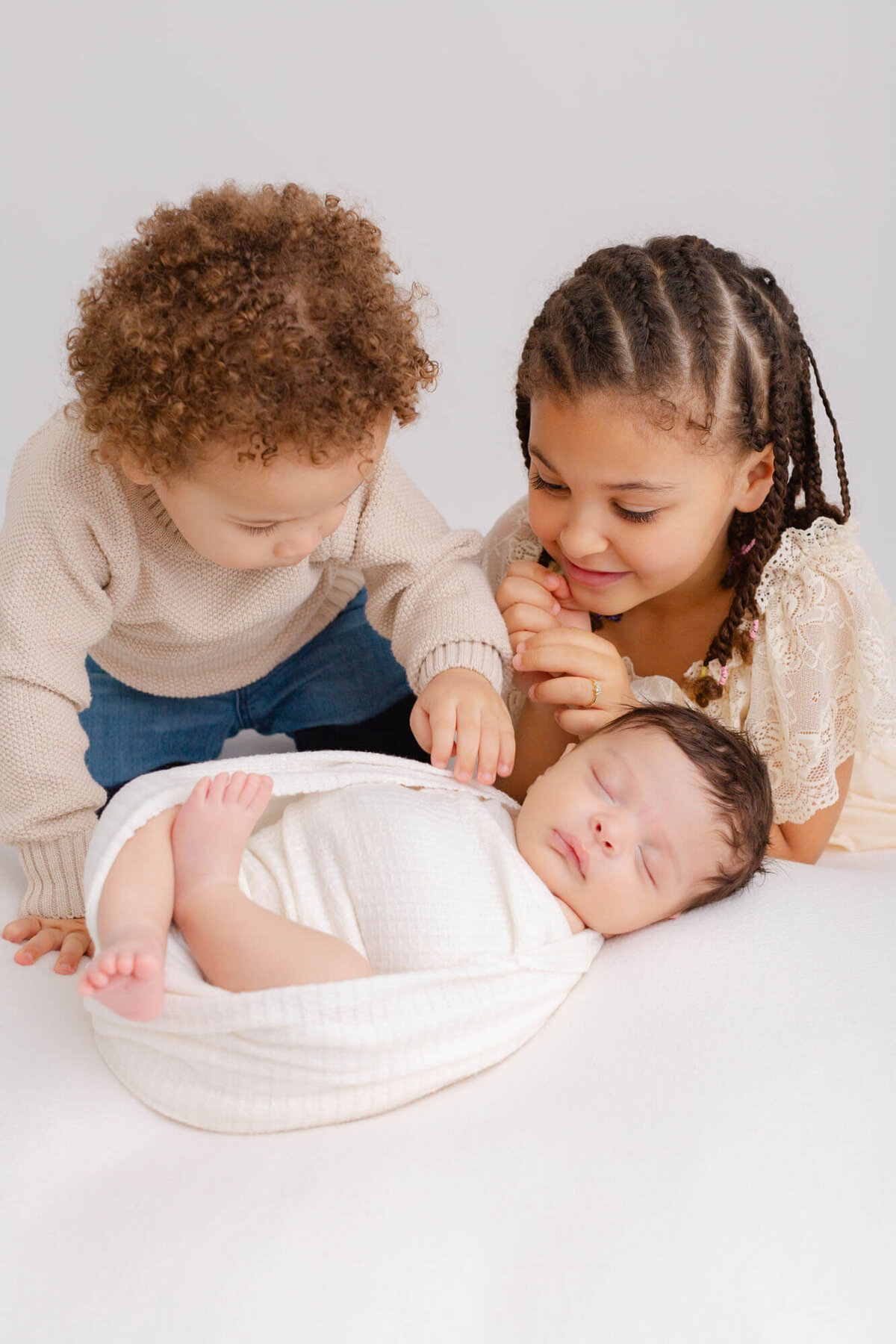 baby sleeping on white blanket while big brother and big sister look down at him.