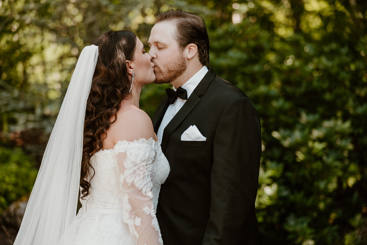 A bride in a lace dress steps up to kiss her groom in a forest