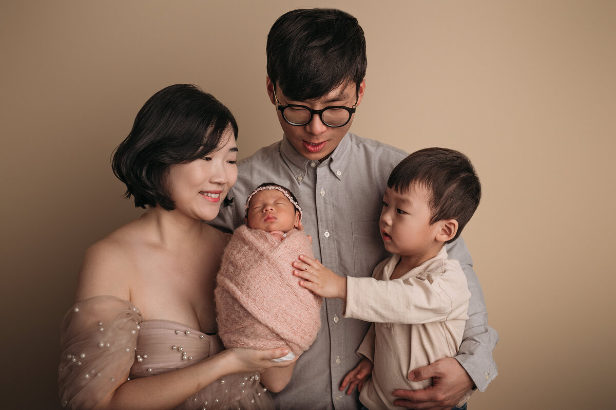 new mom wearing pearl gown  standing next to dad with glasses with  a grey shirt on standing next to their toddler son wearing beige shirt while they all hold and look at their newborn baby girl in front of cream backdrop