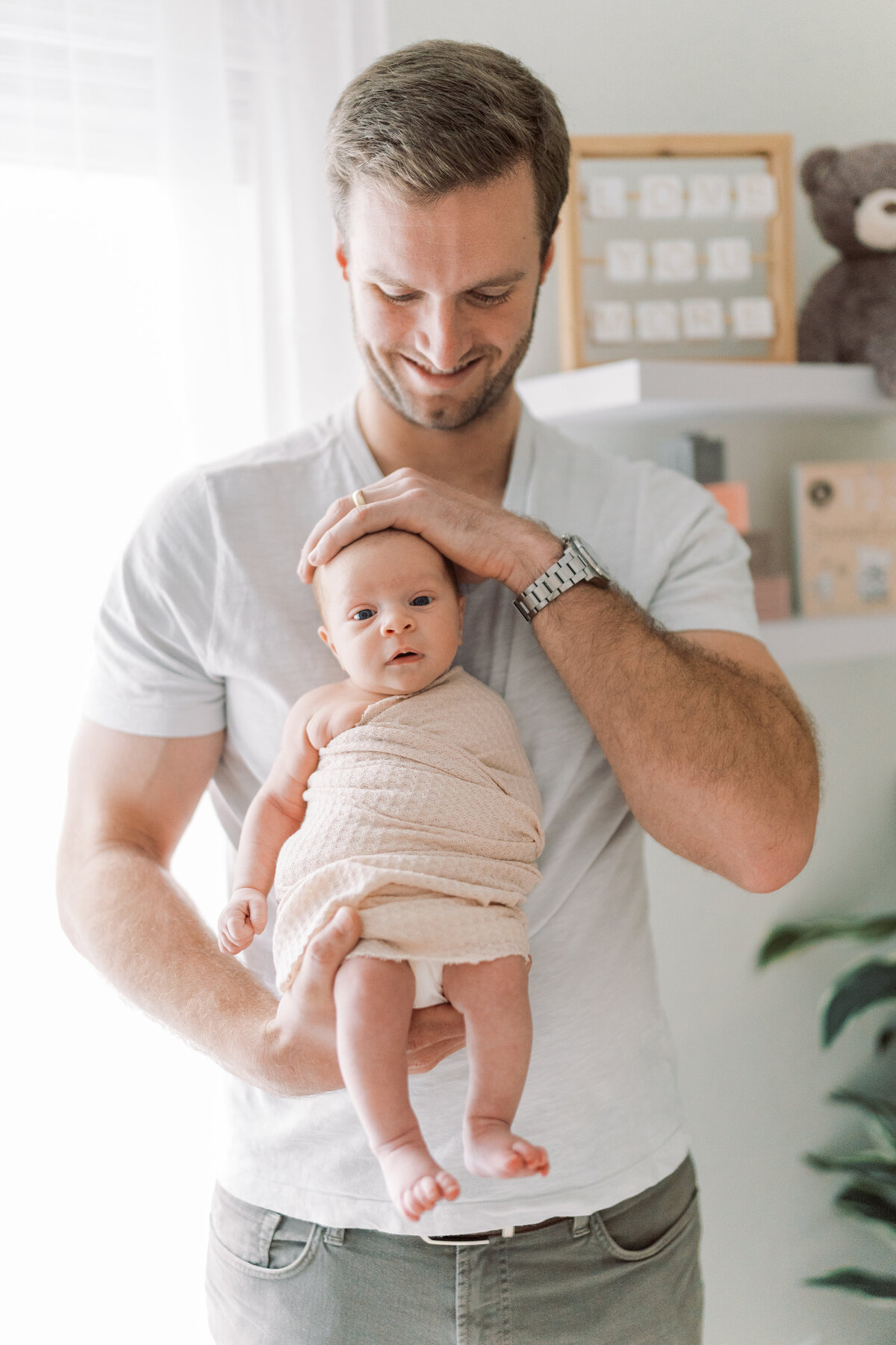 Portrait of a man in a grey shirt and grey jeans holding a swaddled baby in front of his chest in baby’s nursery.