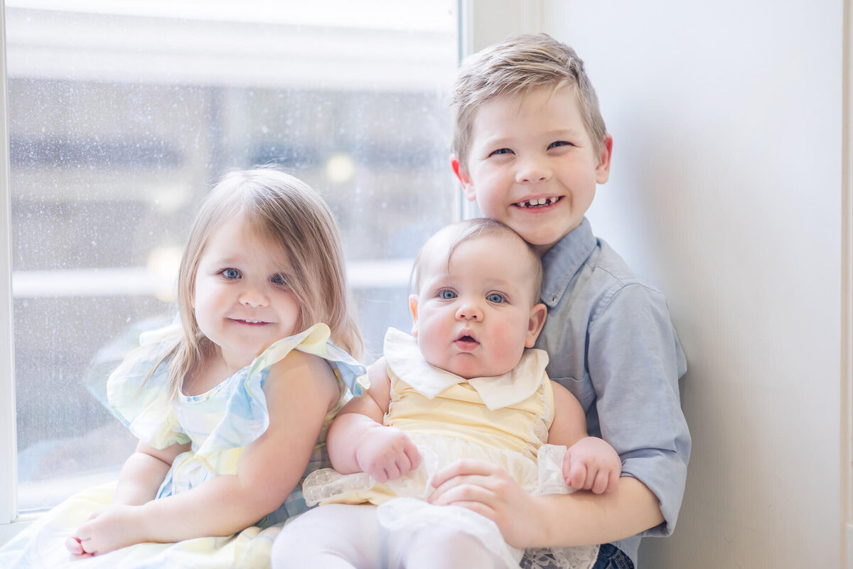 three siblings sitting by a window wearing light blue and pale yellow