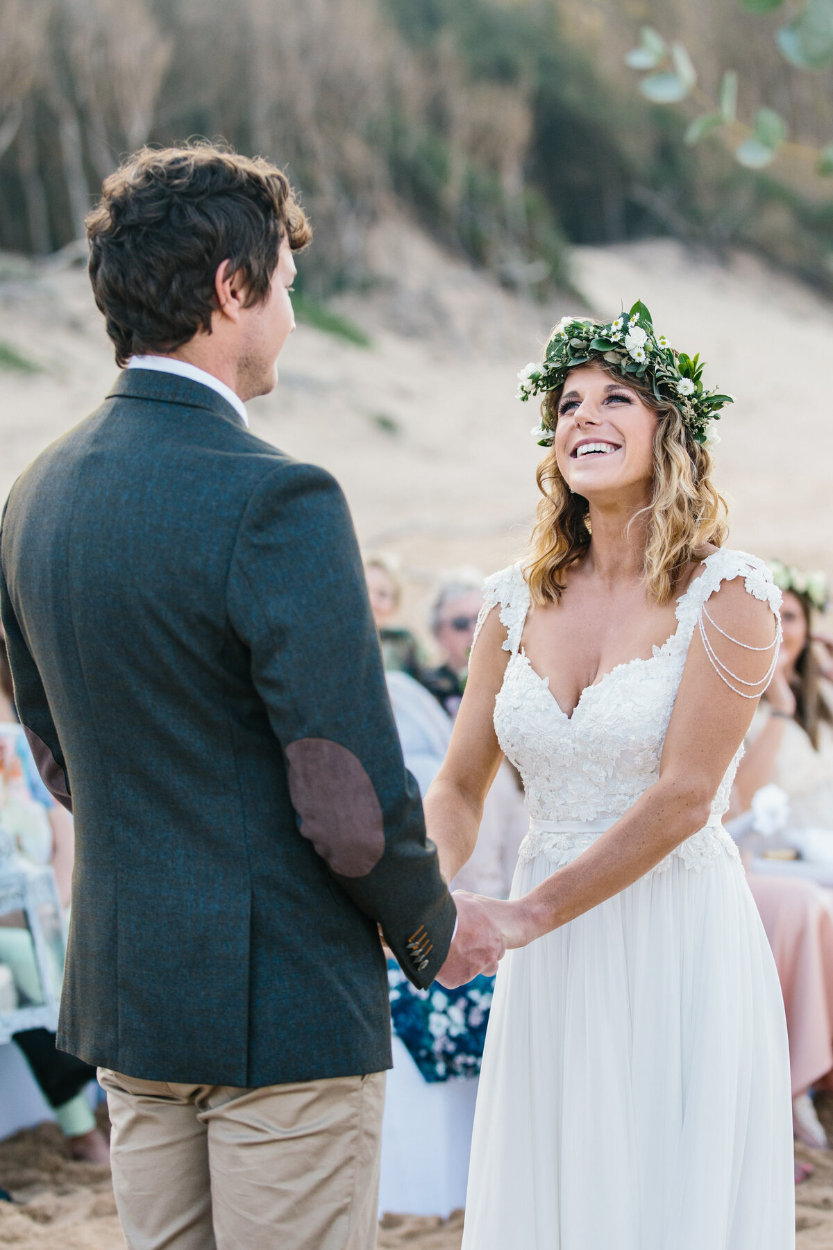 Smiling bride looks into groom's eyes while holding hands on the beach. 