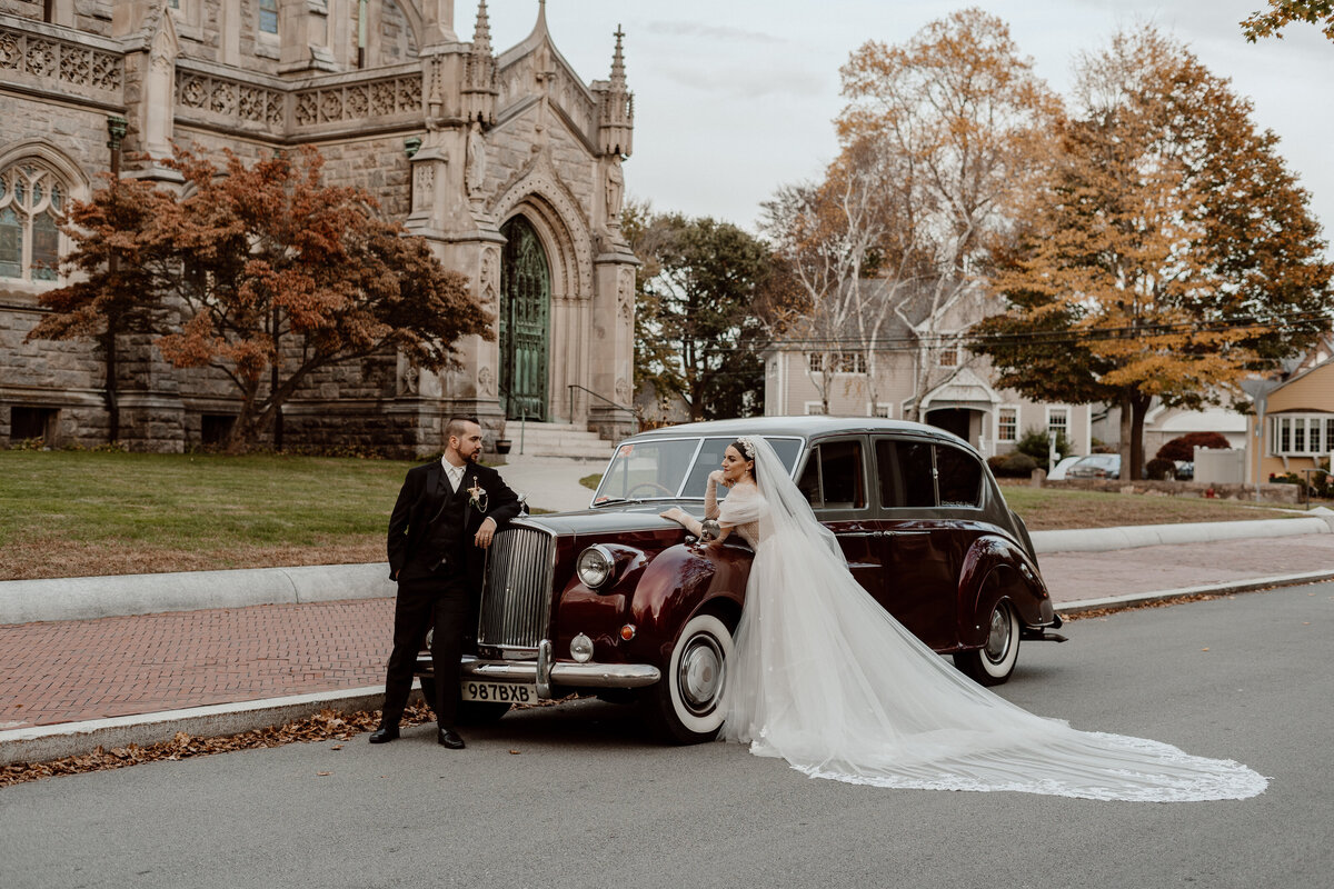 A bride and groom pose with a vintage car in front of an ornate stone church. The bride, wearing a long flowing veil and a white off-the-shoulder gown, leans on the car while smiling at the groom. The groom, dressed in a black suit, stands beside her, looking at her lovingly. The maroon vintage car adds a classic touch to the scene, which is set against the backdrop of the church's gothic architecture and autumnal trees.
