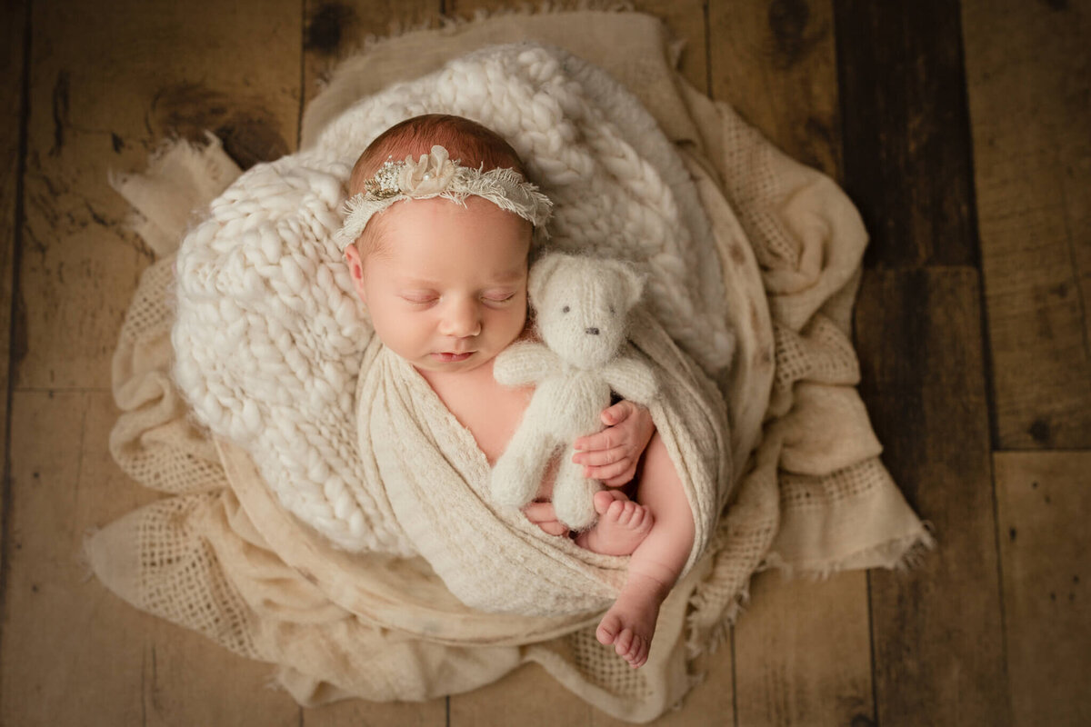 baby girl wrapped up in blanket holding tiny white teddybear on dark wood backdrop.