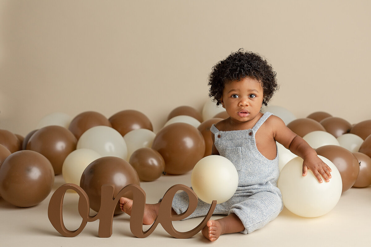 columbus-ohio-first-birthday-cake-smash-photographer-baby-boy-in-light-gray-knit-romper-surrounded-by-cream-and-dark brown-balloons-with-wood-one-sign