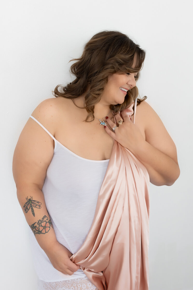 a 40 year old plus size woman smiles happily while holding a pink satin sheet. she is wearing a white tank top of the shoulder strap has fallen down.