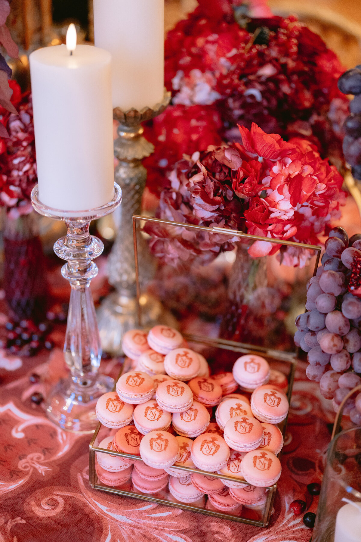 The photo was taken at Shangri La Hotel & Ritz Paris Hotel Paris, France, by photographer Matteo Coltro, with Zen Film Works as Videographers. The exquisite image captures a decorative table setting showcasing pink macarons neatly arranged in a glass box. The table is further adorned with vibrant red and purple flowers, clusters of grapes, and elegant candles in crystal holders, creating a luxurious and inviting ambiance. It truly epitomizes elegance and sophistication.