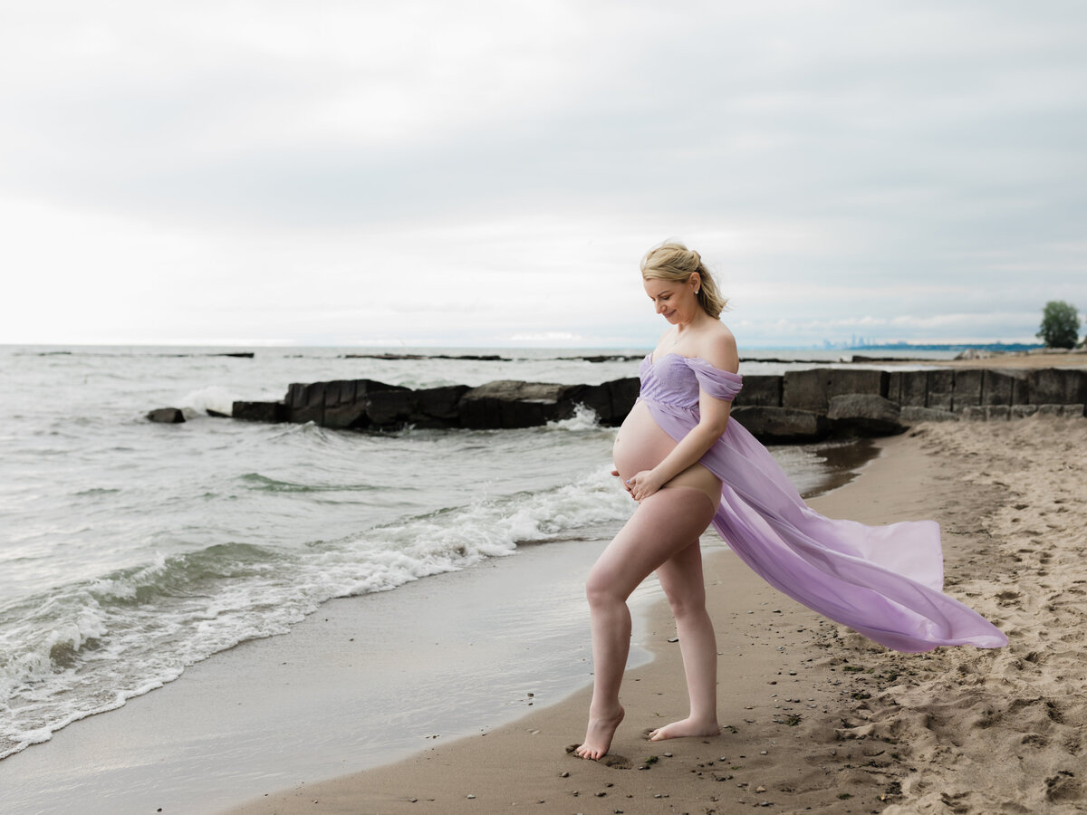 pregnant mother in purple maternity gown holding baby bump on beach for maternity photoshoot 