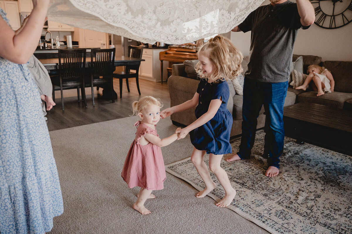 two sisters dance to music in their living room while mom, holding their baby brother and dad shake a blanket up and down above them