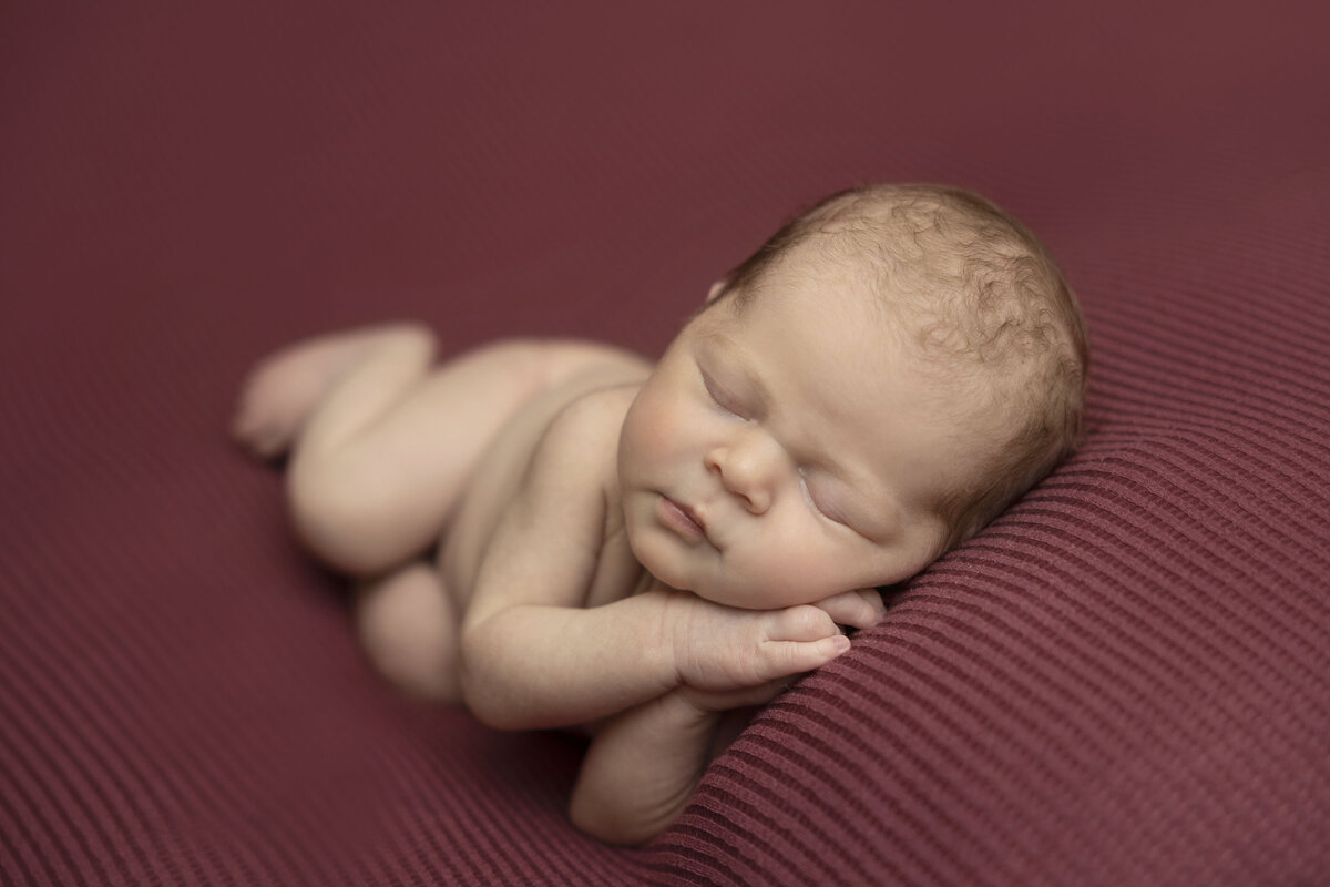 Newborn sleeps with her hands under her cheek on pink striped blanket.