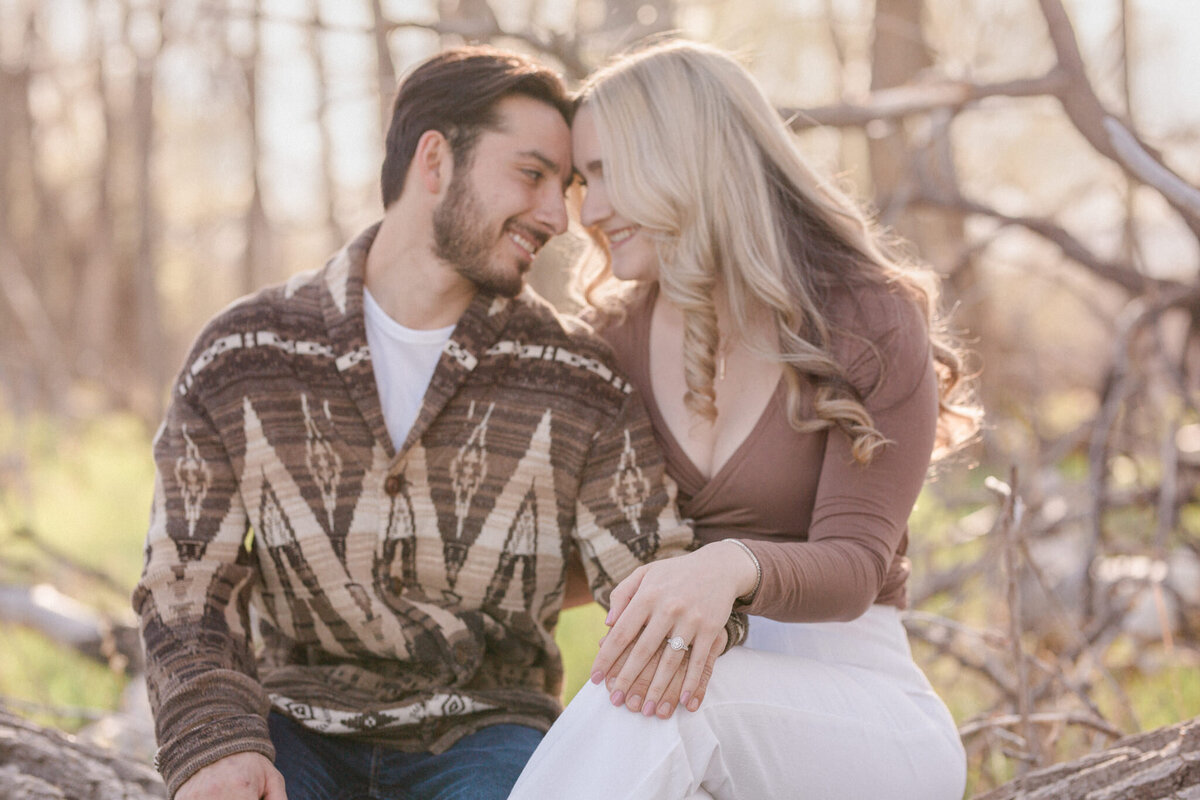 A dark haired man and his tall blond fiancee sit on a fallen tree and show off her new diamond engagement ring  in Boulder, Colorado