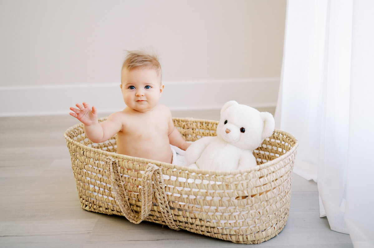 Baby boy sits in moses basket and waves during baby photography session in Raleigh photography studio