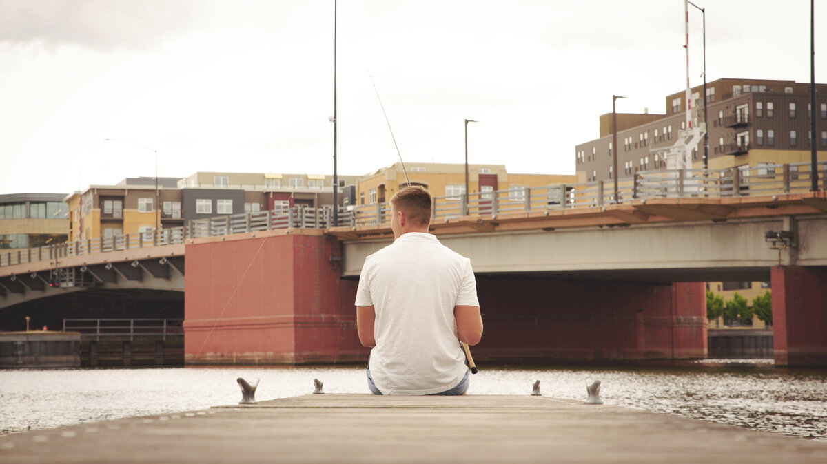 De Pere High School senior boy wearing blue shorts and a cream hooded sweatshirt sitting on dock by The Depot in Downtown Green Bay, Wisconsin