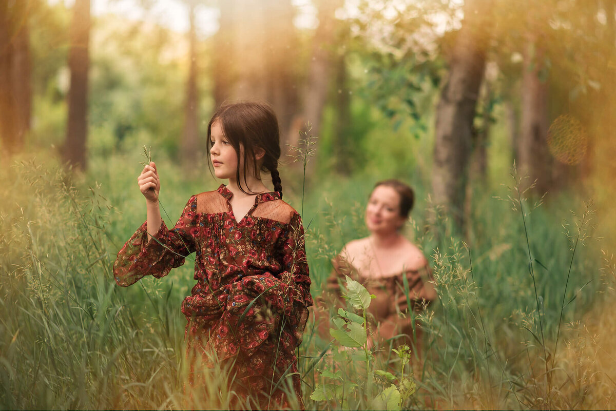 Mother sits in the grass back in the distance while looking at her daughter in the foreground. Daughter is standing playing with grass in her hands. They both smile. taken by brittany danielle