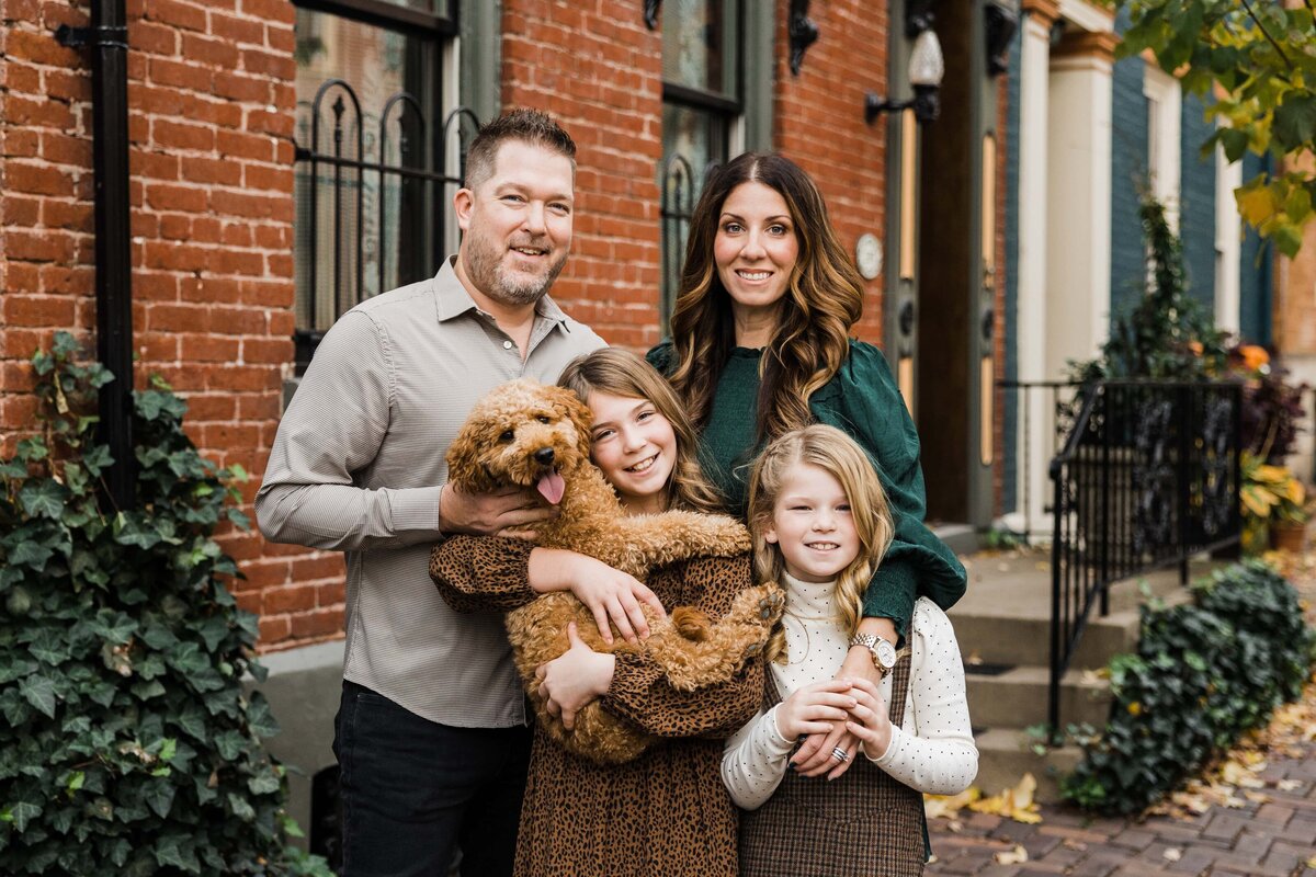 A family of four with their dog smiling for a photo in a quaint, brick-lined street setting, captured by a skilled Pittsburgh family photographer.