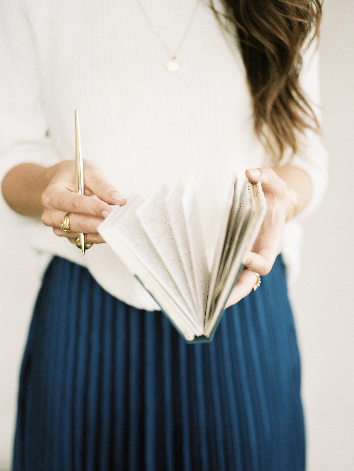 woman in cream sweater, blue pleated skirt, gold jewelry with journal, gold pen