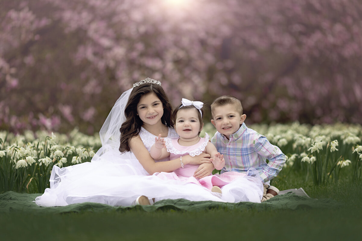 A young girl in a large white dress and tiara sits on a blanket with her toddler brother and sister in a field of white daffodils for a New Jersey Communion Portrait Photographer
