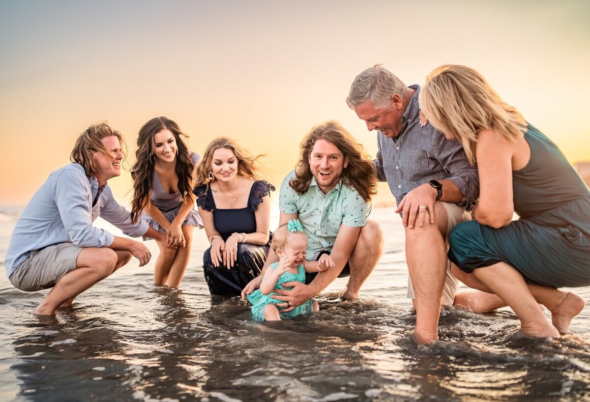 Multi-generational family gathered around a baby in the ocean at sunset, laughing and splashing together
