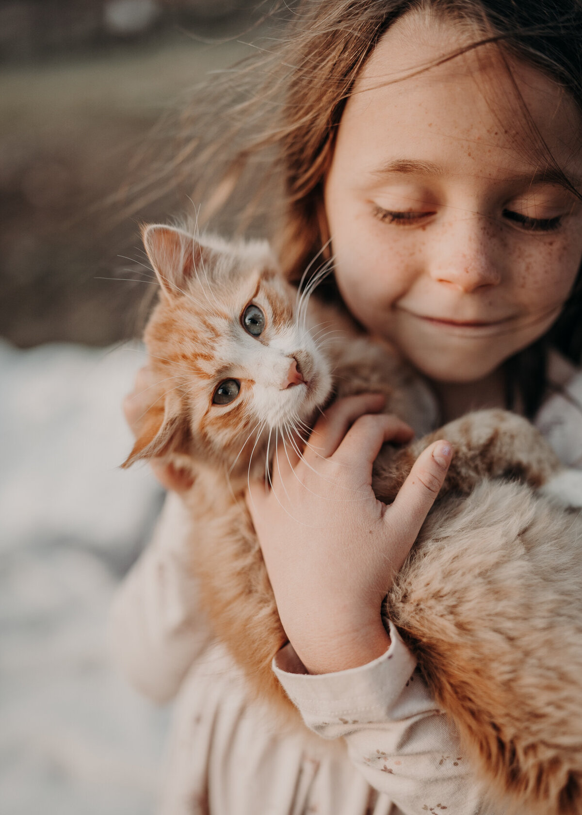 A young girl looks out of the corner of her eye at her adorable tan barn cat that stares directly at the camera, while the wind blows her hair during a golden our family photo session in the outskirts of Denver, Colorado.