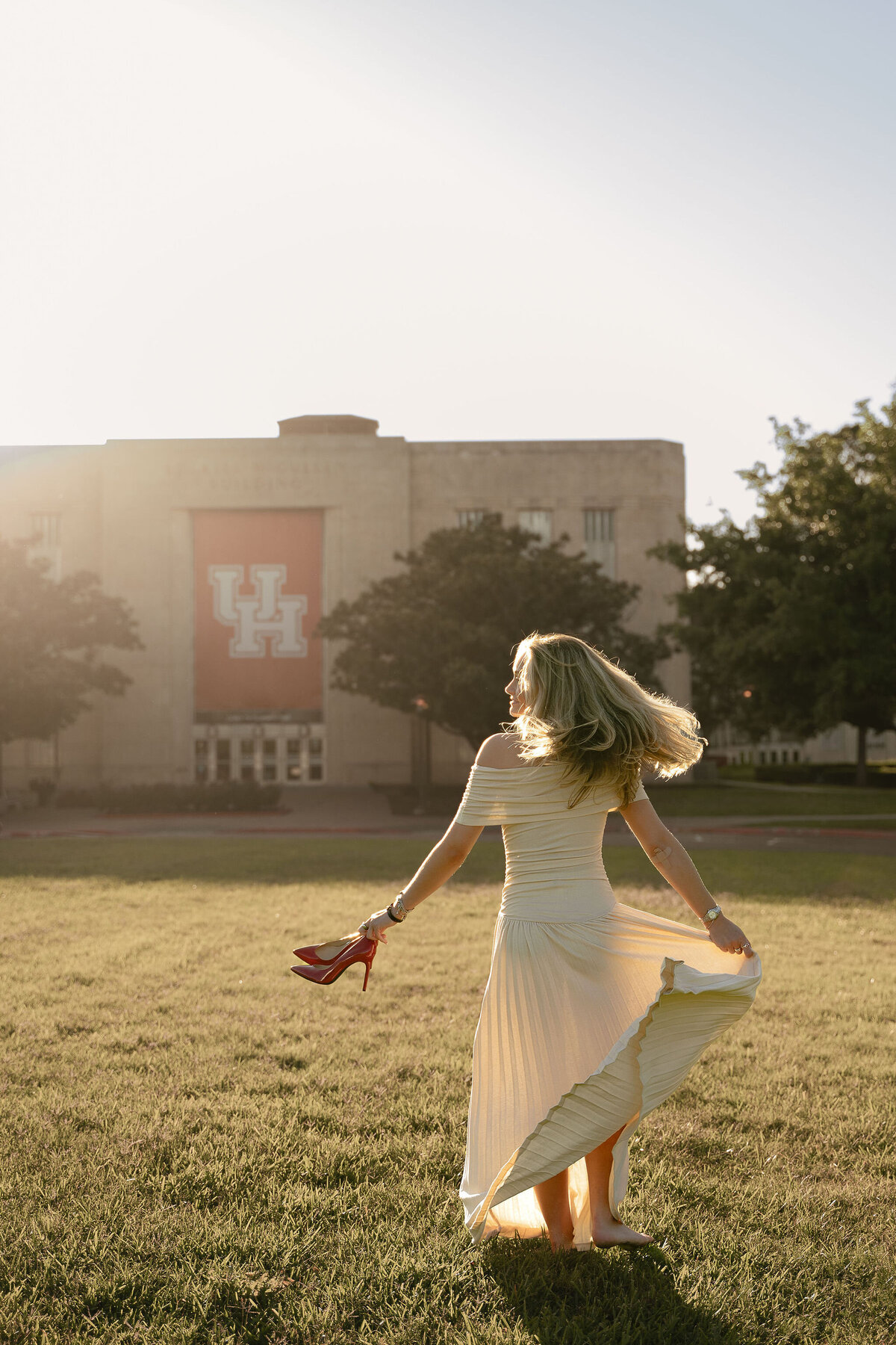 A woman in a flowing white dress walks barefoot on grass, holding red shoes. The sun sets behind a large building with "UH" on a banner, creating a serene and nostalgic mood.