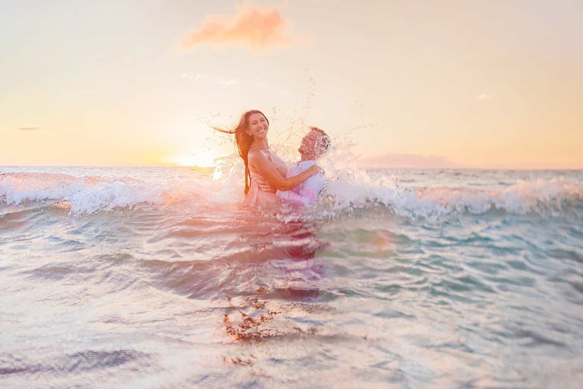 Couple in the ocean at sunset on Maui get splashed by a wave as Love + Water photograph their engagement session from the beach