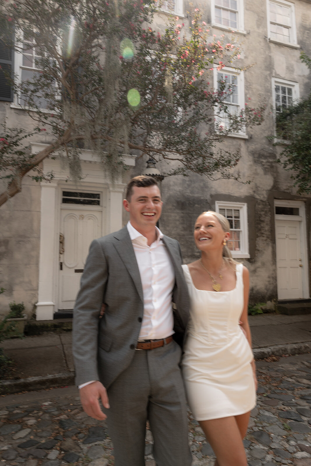 Man in grey suit and woman in white short dress walking on cobblestone street in Charleston for enagagement photos