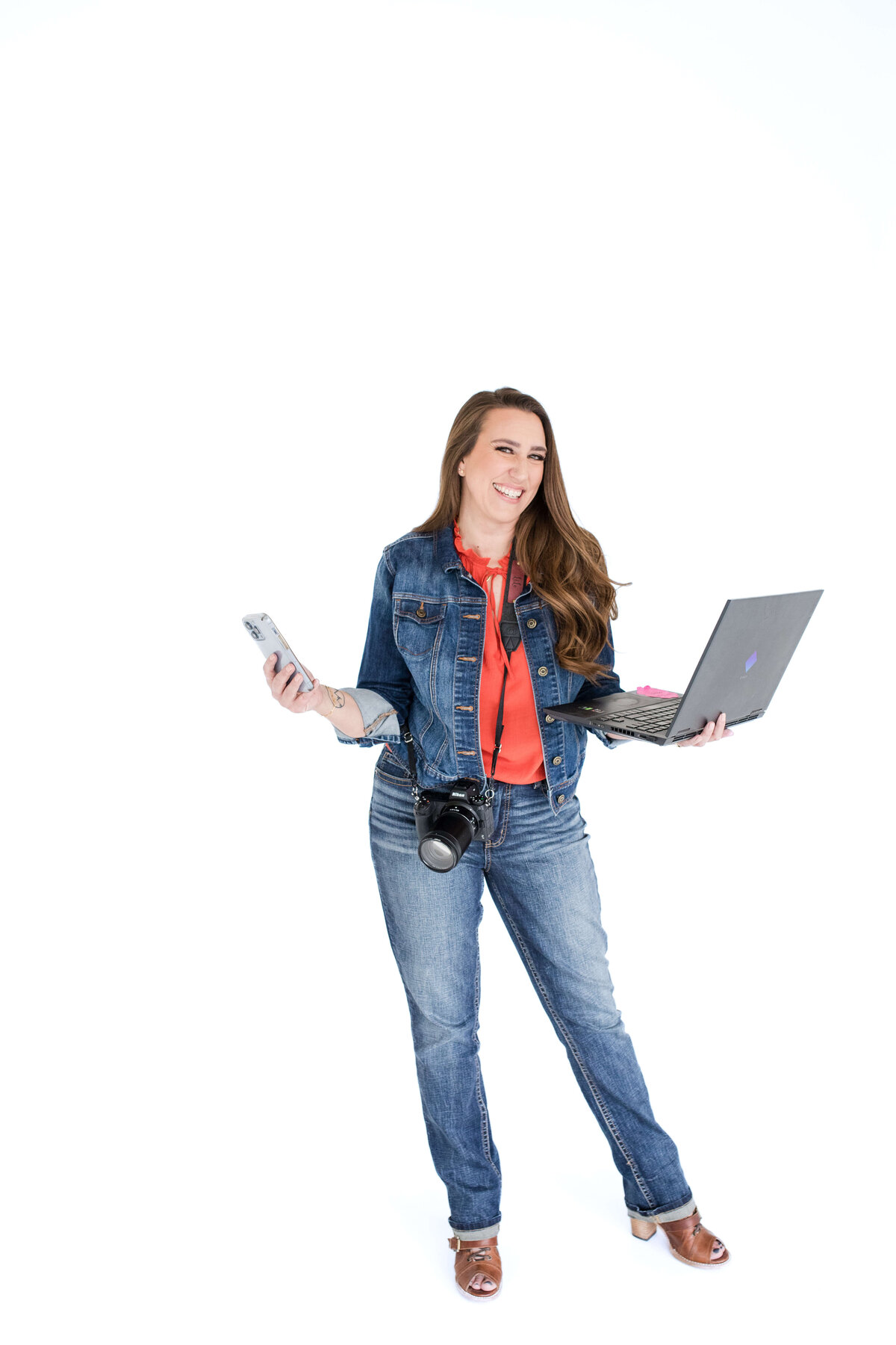 brand photo of photographer holding a laptop and a camera and laughing captured by brand photographer near me at a studio session with a white backdrop