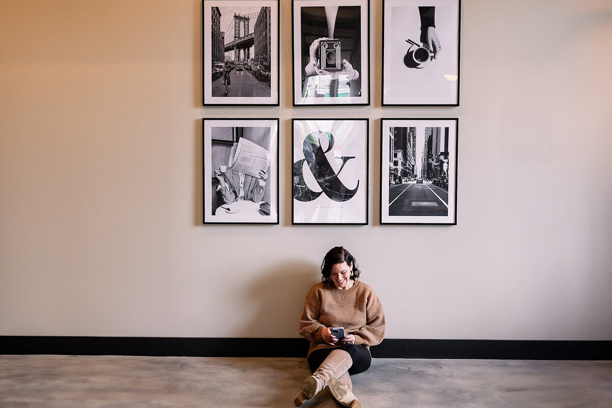 Woman sitting on the ground while looking at her phone underneath photo frames