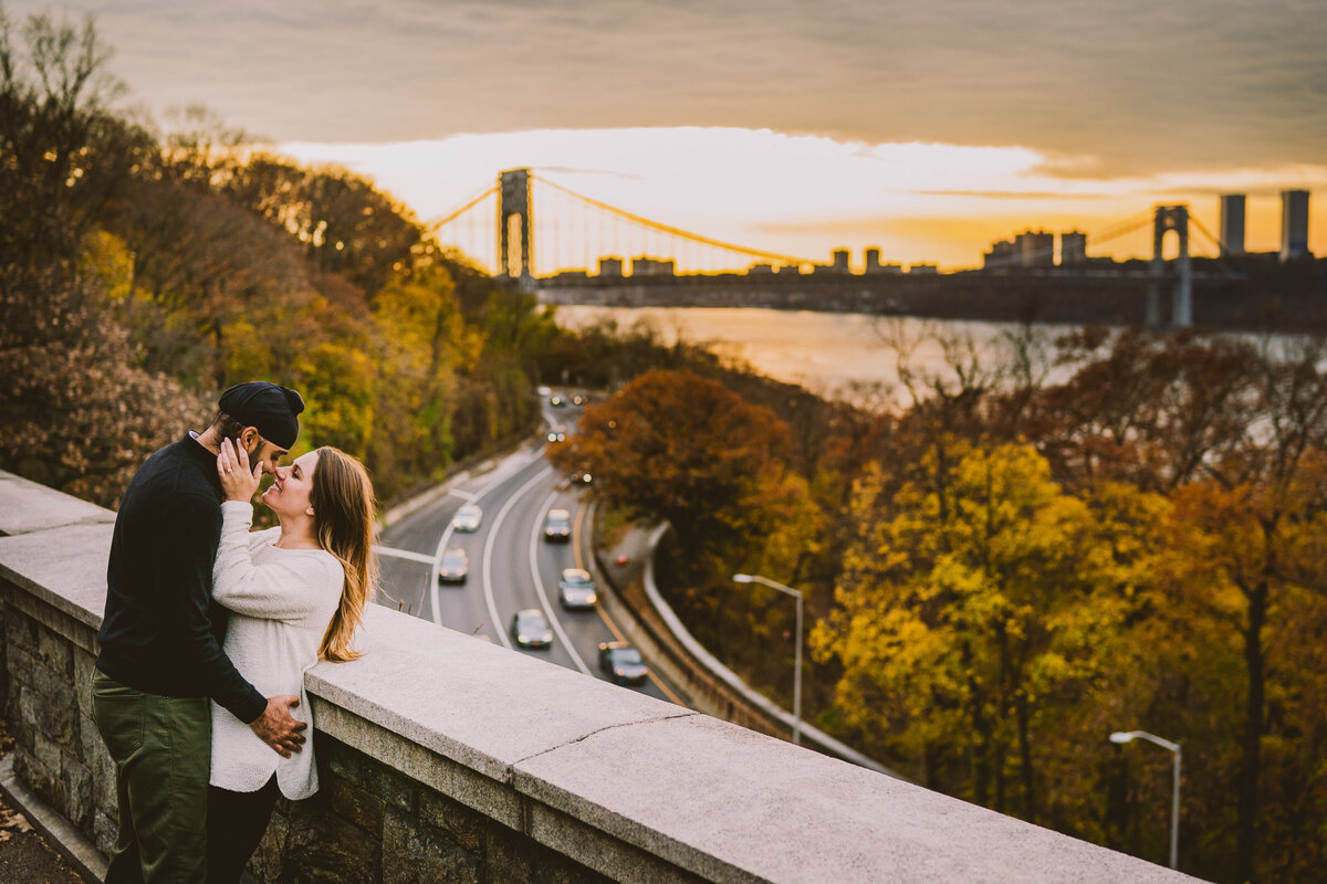 An engaged couple share a moment near the ledge at fort tryon park with the view of the west side highway and bridge