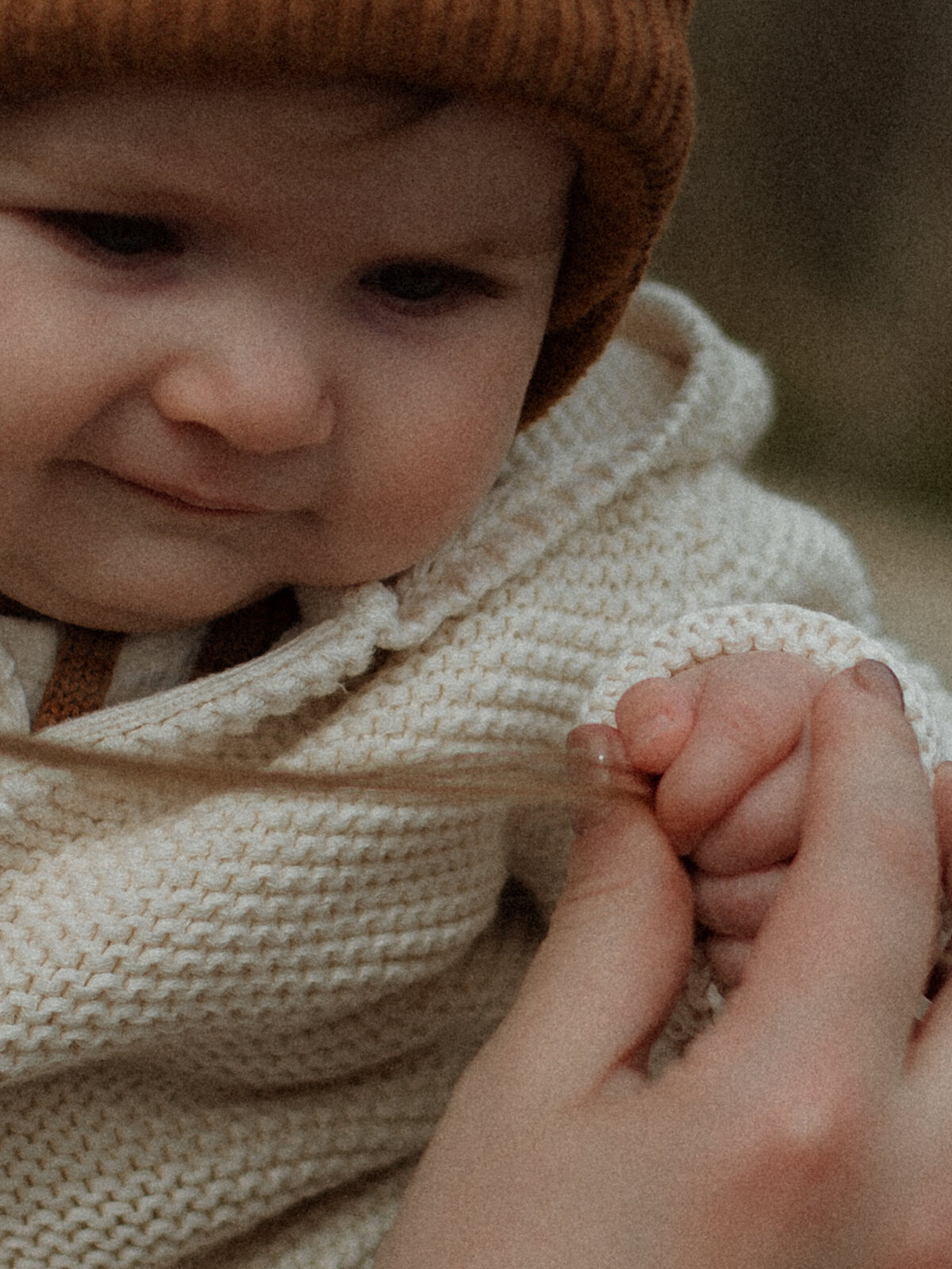 A close-up of a baby’s face wearing a knit hat and sweater, gripping mom’s hair with a soft and curious expression.