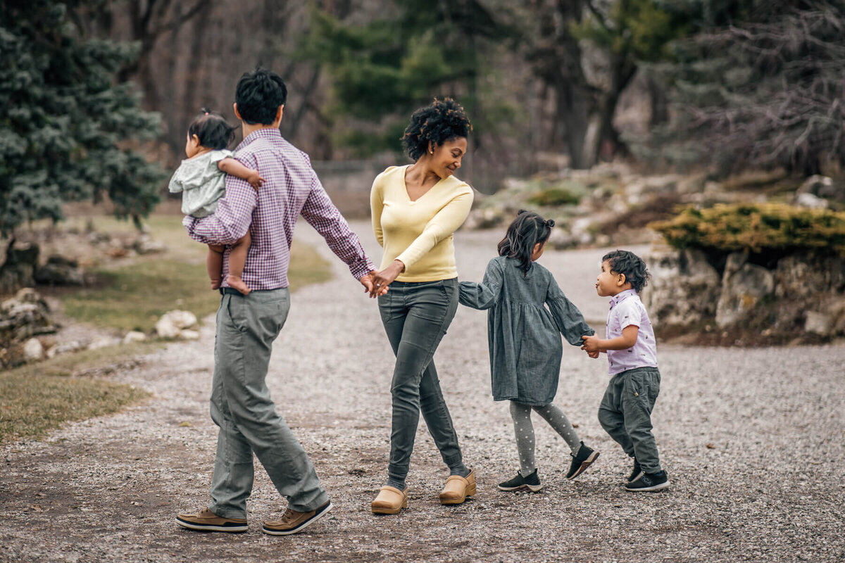 A family joins hands and walks in a line, looking back at each other during a fun filled family photoshoot at the Lake Harriet Peace Gardens in Minneapolis, MN.