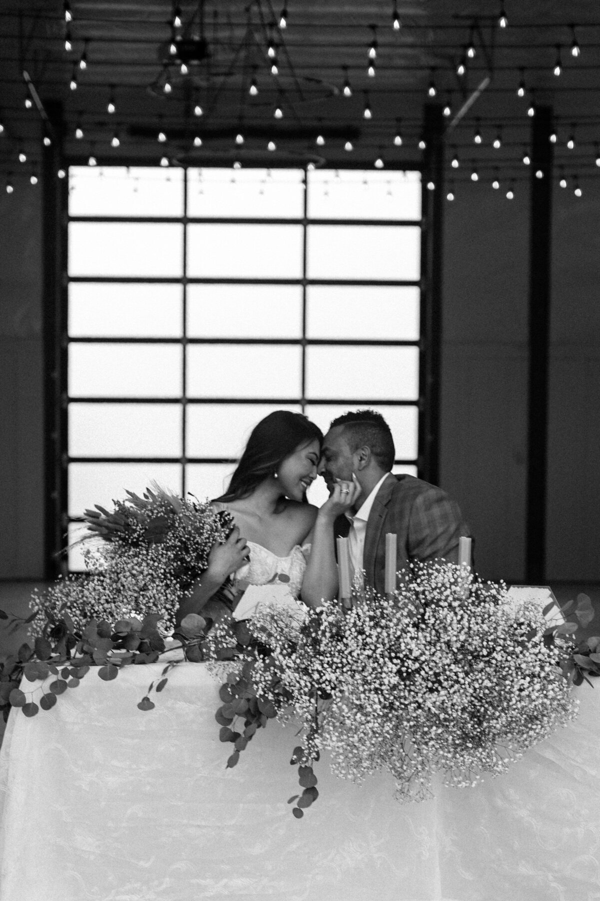 B&W photo. Couple touching foreheads in front of beautiful reception table with big window behind them.