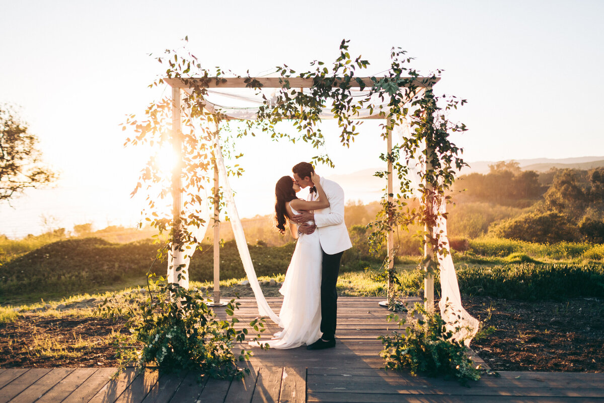 A colorful photograph of Sarah and Erik on their wedding day at Dos Pueblos Orchid Farm in Santa Barbara, California. The bride and groom are under a chuppah adorned with greenery and white flowing fabric. The portrait is taken at sunset with the sun glowing behind them, lighting up the view of the green rolling seaside hills. The bride and groom are standing under the chuppah in an embrace with his arms around her waist and her hand on his neck as they kiss. He is in a tuxedo with white jacket and black pants and she is in a white dress with a low back that is bustled. Wedding photograph by Stacie McChesney of Vitae Weddings.