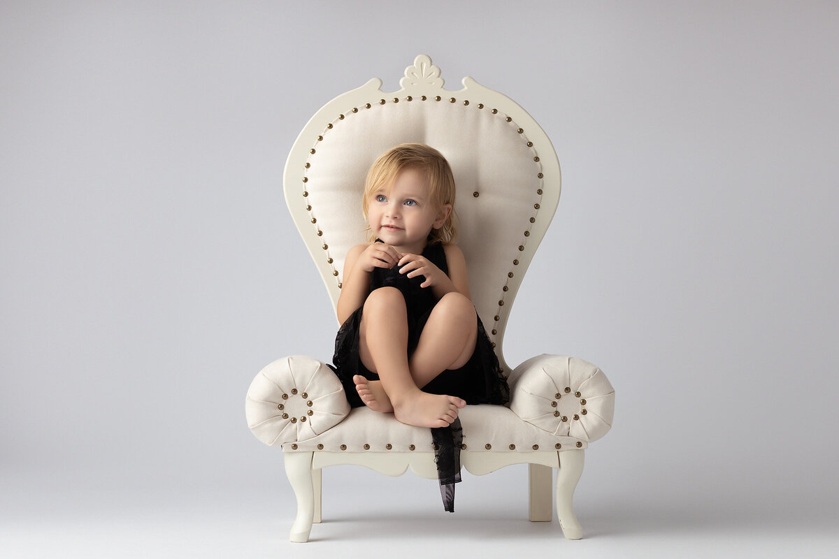 Toddler posing on a white chair in a black dress, captured by Dallas family photographer.